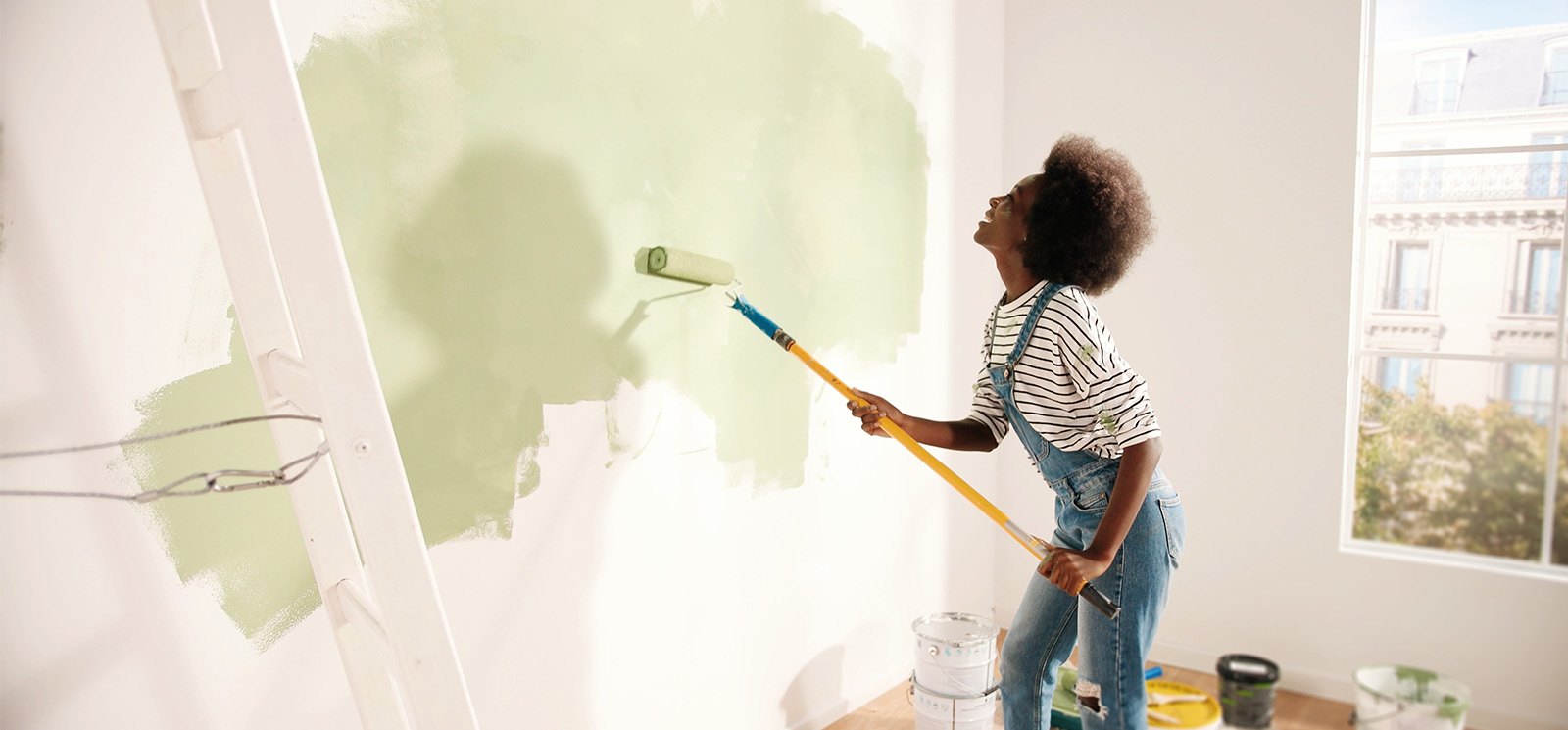 Young African American woman dancing and painting wall with roller brush while renovating apartment.