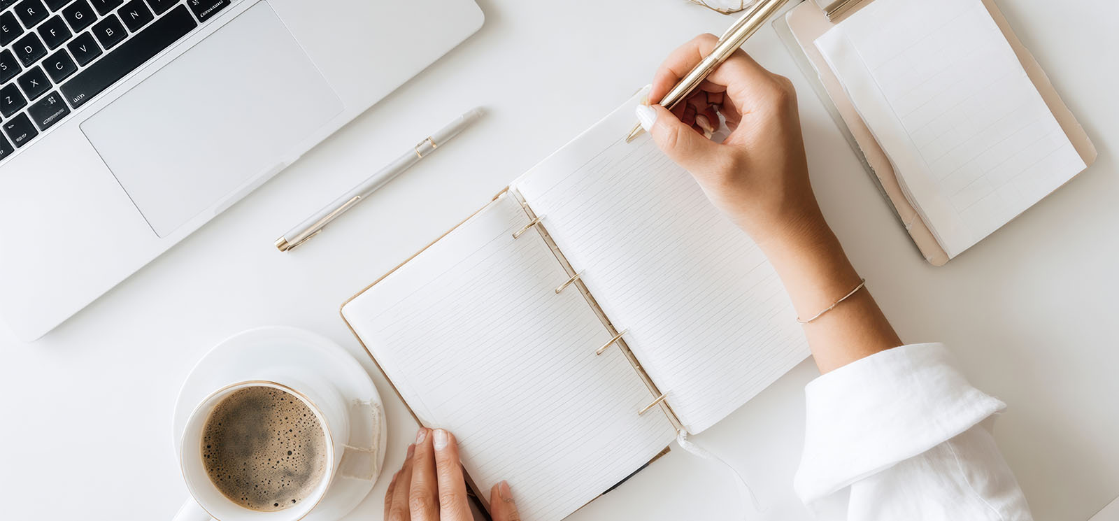 Person writing goals in planner on desk with laptop and coffee.