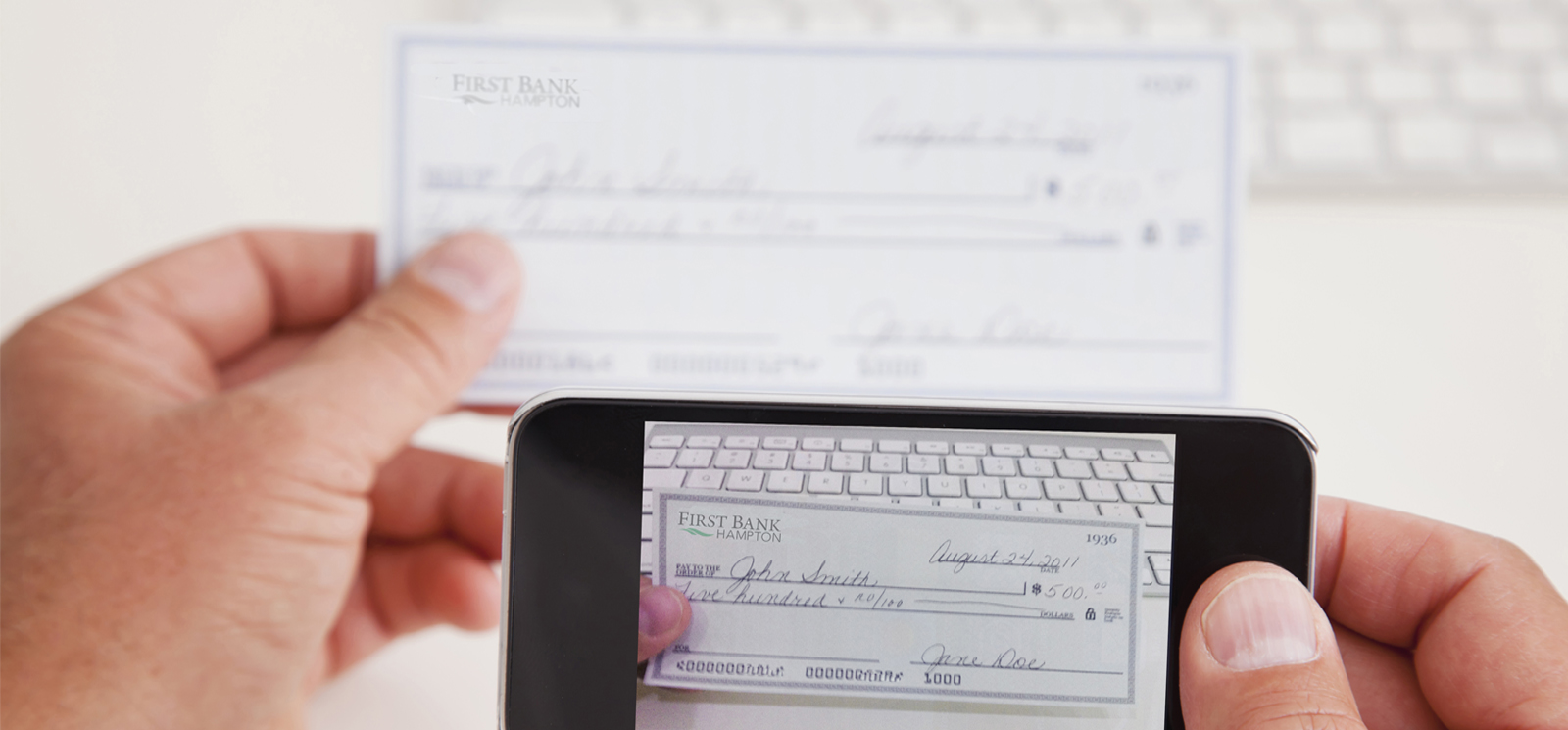 Shows a hand holding a smart phone with the image of a check to be deposited. The check is being held in the background by the other hand.